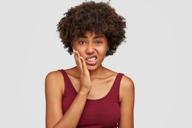 Displeased beautiful african american woman keeps hand on cheeks, feels toothache, clenches teeth from pain, has dark healthy skin and crisp hair, dressed casually. negative facial expressions Free Photo