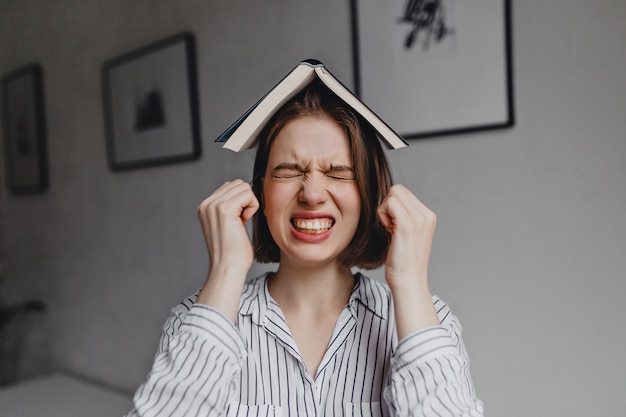 Displeased girl in striped shirt is angry with her eyes closed and open book on her head against background of room with pictures. Free Photo