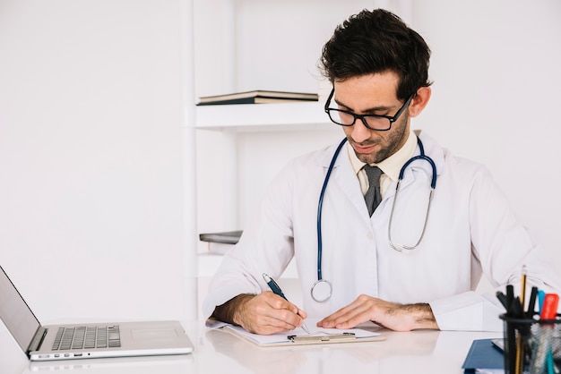 Doctor writing on clipboard with laptop on desk | Free Photo