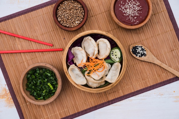Dumpling with salad in bamboo steamer surrounded with chives; coriander seeds and chopsticks on placemat Free Photo