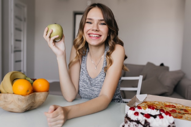 青リンゴとオレンジで朝食を楽しんでいる恍惚とした若い女性 果物やケーキを食べるポジティブな白人の女の子の屋内写真 無料の写真