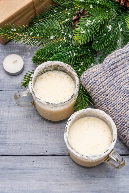 Eggnog cocktail in two glasses mug arranged with christmas decoration on light wooden table. evergreen fir tree branch, gift, cozy jersey, artificial snow Premium Photo