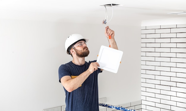 Electrician builder with beard worker in a white helmet at work, installation of lamps at height. professional in overalls with a drill on the repair site. Free Photo