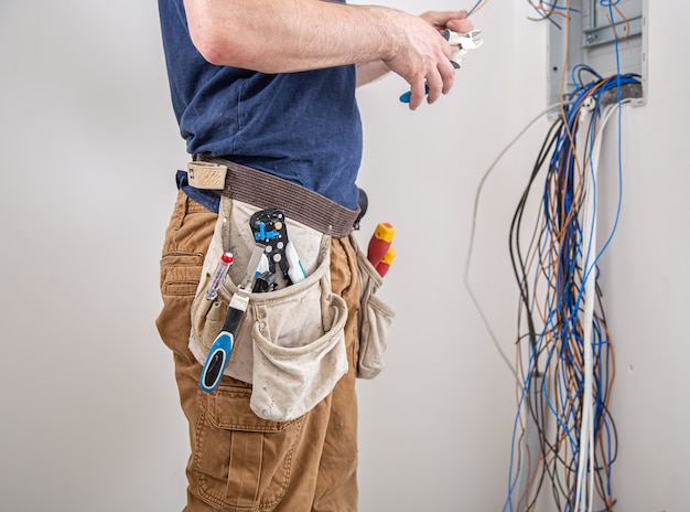 Electrician builder at work, examines the cable connection in the electrical line in the fuselage of an industrial switchboard. professional in overalls with an electrician's tool. Free Photo