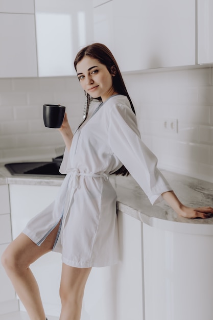 早朝のコーヒーを飲みながらキッチンの前に立っている白いローブでエレガントな笑顔の女性 無料の写真