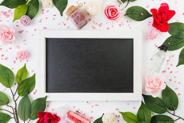 Elevated view of blank frame surrounded with cosmetic products and leaves on white surface Free Photo