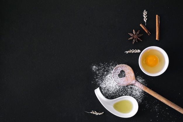 Elevated view of egg; spices; grain; heart shape spoon; flour and oil on black surface Free Photo