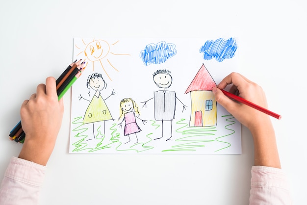 An elevated view of a girl's hand drawing the family and house with colored pencil on drawing paper Free Photo