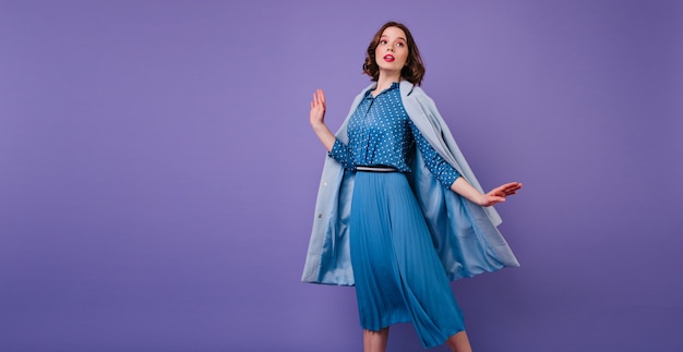 Emotional brunette woman in blue coat posing on purple wall . indoor photo of beautiful short-haired female model in trendy midi dress. Free Photo