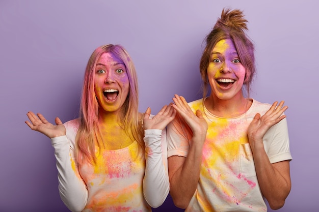 Emtional happy best female friends use coloured dye during holi festival, smeared with colourful rainbow powder, spread palms from happiness and fun, celebrate indian spring holiday, drench each other Free Photo