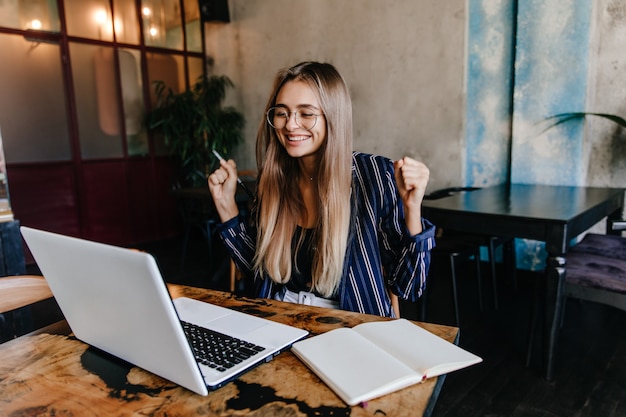 Excited long-haired girl having fun during work with computer. indoor photo of smiling female freelancer using laptop in cozy cafe. Free Photo