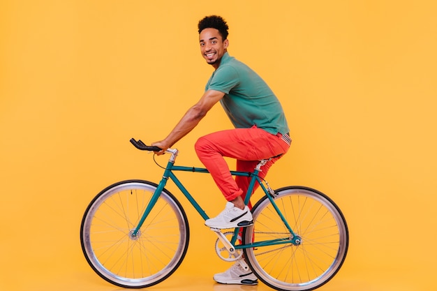 自転車でポーズをとるスポーツの白い靴で興奮した男性モデル 自転車に座って見ている陽気なアフリカ人の屋内写真 無料の写真