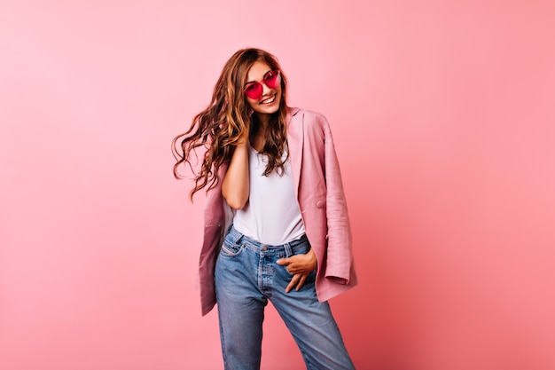 Excited white girl in bright stylish glasses posing on pink. dreamy curly woman playing with her ginger hair and laughing. Free Photo