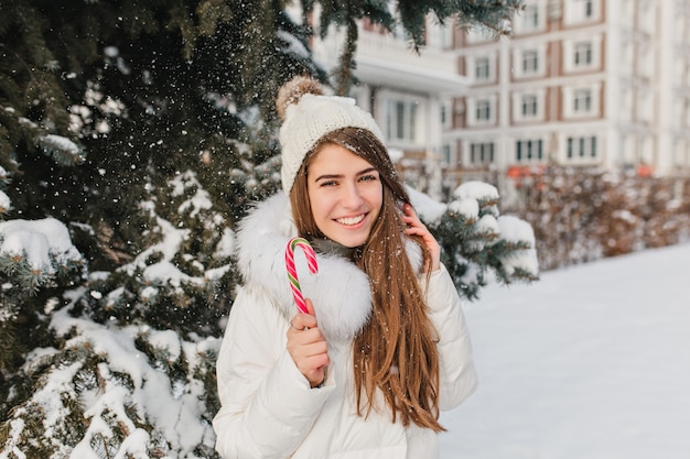 雪の日に楽しんで写真撮影を楽しんでいるストレートの茶色の髪の興奮した女性 クリスマスのお菓子でポーズ流行の服で見事な白人女性の屋外のポートレート 無料の写真