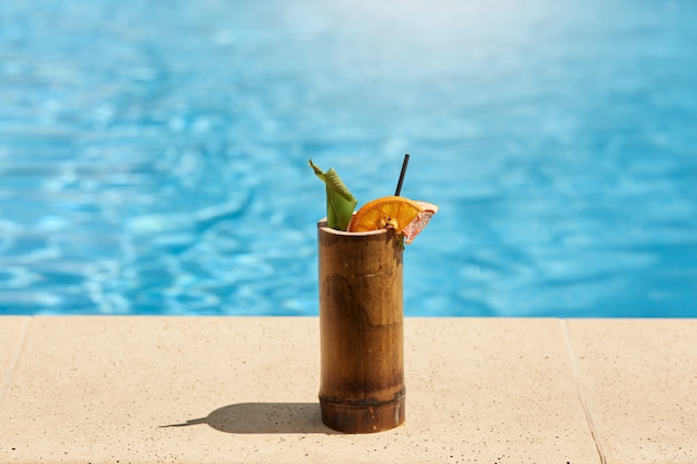 Premium Photo Exotic Cocktail In Wooden Glass With Lemon And Drinking Tube Standing On Poolside With Blue Water On Background