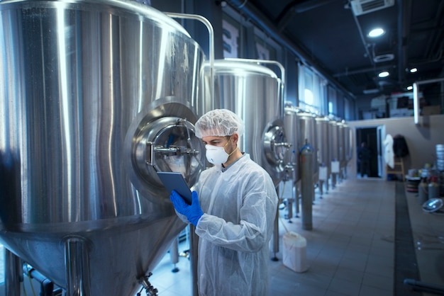 Factory worker technologist in protective uniform with hairnet and mask controlling food production on tablet computer Free Photo