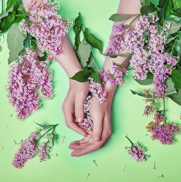 ファッションアートは 自然化粧品の女性 明るいコントラストのメイク ハンドケアと手に明るい紫色のライラックの花を手渡します 対照的な緑の背景にテーブルに座っている女の子の創造的な美しさの写真 プレミアム写真