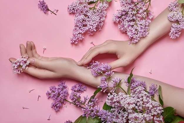 ファッションアートは 自然化粧品の女性 明るいコントラストのメイク ハンドケアと手に明るい紫色のライラックの花を手渡します 対照的なピンクの背景にテーブルに座っている女の子の創造的な美しさの写真 プレミアム写真