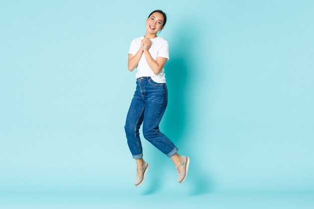 Fashion, beauty and lifestyle concept. happy and dreamy, excited asian girl in casual outfit, jumping from happiness and joy, clap hands enthusiastic, standing over light blue wall Free Photo
