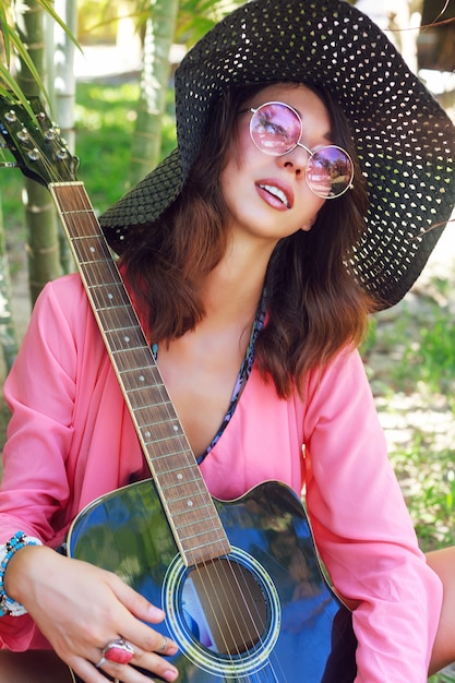 自然なメイクアップとギターで庭でポーズをとってふわふわのブルネットの毛の美しい少女のファッションの肖像画 帽子と丸いトレンディなピンクのサングラス 無料の写真