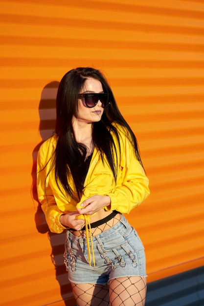 スタイリッシュな若者の黄色のジャケット デニムのショートパンツ オレンジ色の都市壁の背景にポーズのサングラスを着ておしゃれなかわいい若い女性 プレミアム写真