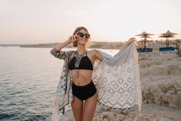 夕暮れ時のビーチでポーズおしゃれな美しい女の子 黒いビキニ ハイウエストの水着 カーディガン 装飾品のケープ 美しい海 岩を身に着けているセクシーなスリムな女性 無料の写真