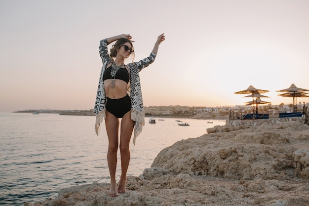 ビーチ 日没 休暇の女の子でポーズをとっておしゃれなモデル 黒のビキニ ハイウエストの水着 カーディガン 装飾品のケープを着ているセクシーな女性 無料の写真