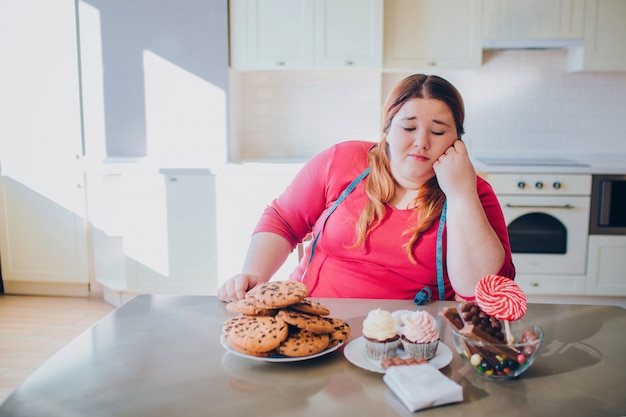 Få Evaluering Berettigelse Premium Photo | Fat young woman in kitchen sitting and eating sweet food.  bored plus size model look at pancakes and sweets on table.