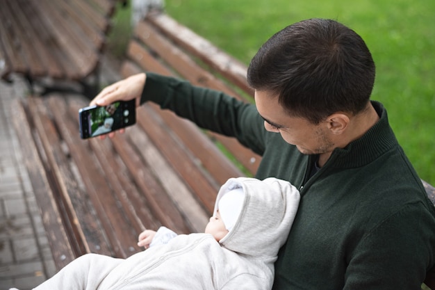 スマートフォンでselfieを取っているベンチに座っている彼の腕の赤ちゃんと緑のジャンパーの父 プレミアム写真