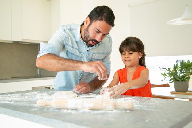 Father teaching his girl to bake bread or pies. focused dad and daughter kneading dough on kitchen table with flour messy. family cooking concept Free Photo