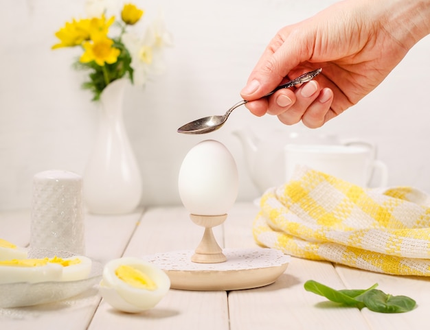 女性の手は白い木製の背景の上のスタンドにスプーンでゆで卵を粉砕します ライトキーの食べ物の写真 プレミアム写真
