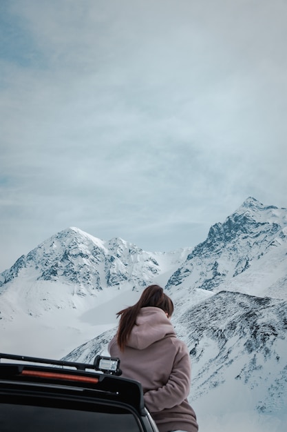 Free Photo Female Leaning On A Black Vehicle In Front Of Amazing Snowy And Rocky Mountains And Cloudy Skies