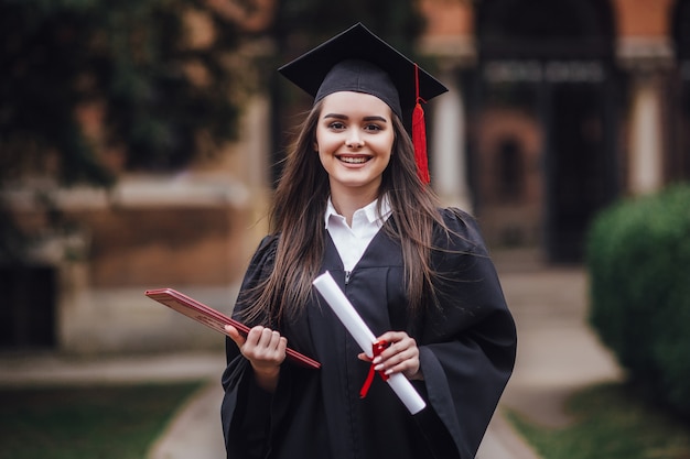 Female student graduate is standing in university hall in mantle, smiling with diploma on hands. Premium Photo