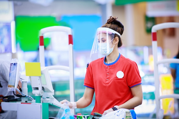 Female supermarket cashier in medical protective mask and face shield working at supermarket. coronavirus concept Premium Photo