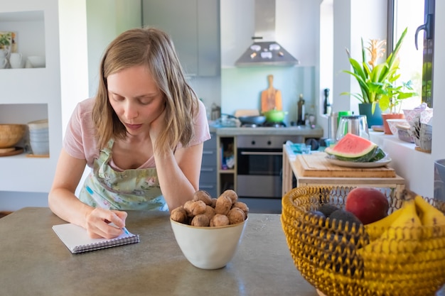 Focused housewife planning weekly menu in her kitchen, writing down grocery list in notebook. cooking at home concept Free Photo
