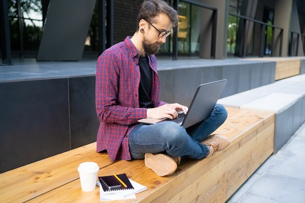 ノートパソコンと木製のベンチにあぐらをかいて座って焦点を当てた男 無料の写真