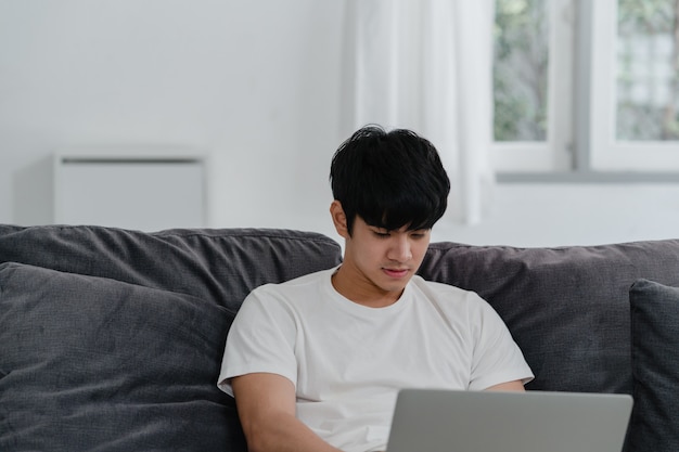 Freelance asian man working at home, male creative on laptop on sofa in living room. business young man owner entrepreneur, play computer, checking social media in workplace at modern house . Free Photo