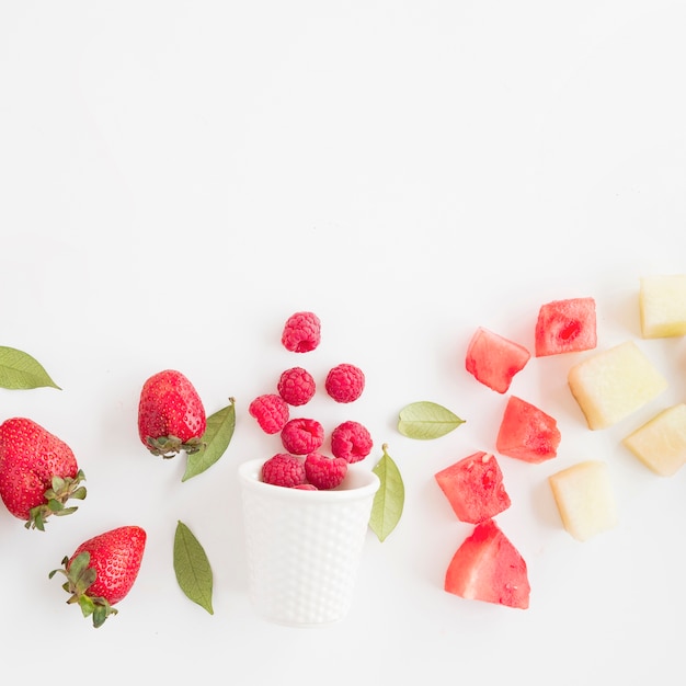 Fresh raspberries spilled front glass with strawberry; watermelon and pineapple isolated on white background Free Photo