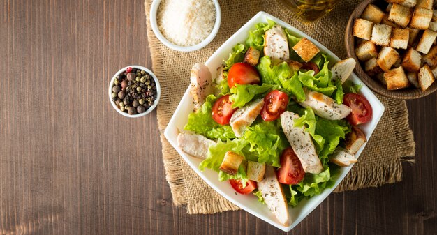 Fresh salad made of tomato, ruccola, chicken breast, eggs, arugula, crackers and spices. caesar salad in a white, transparent bowl on wooden background Premium Photo
