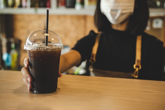 Friendly woman barista wearing protection face mask waiting for serving ice black coffee to customer in cafe coffee shop, cafe restaurant, service mind, small business owner, food and drink concept Premium Photo