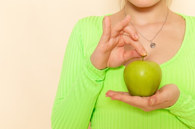 クリーム色の壁のフルーツモデルの女性に緑の新鮮なリンゴを保持している緑のシャツの正面の若い美しい女性 無料の写真