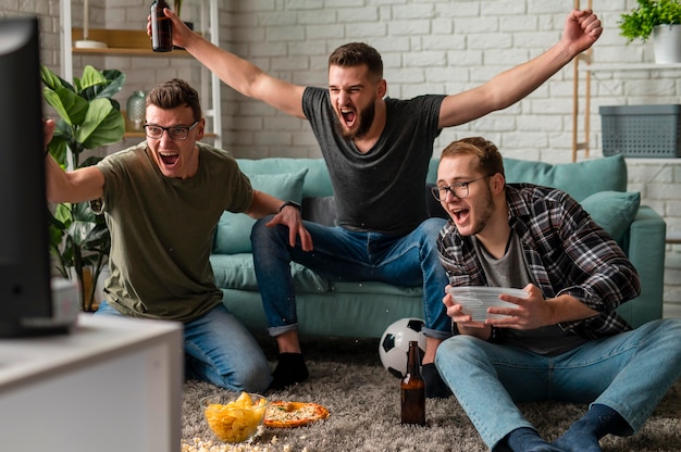 Front view of cheerful male friends watching sports on tv together while having snacks and beer Free Photo