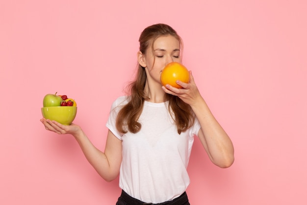 ピンクの壁に新鮮な果物とグレープフルーツの香りがする白いtシャツ保持プレートの若い女性の正面図 無料の写真