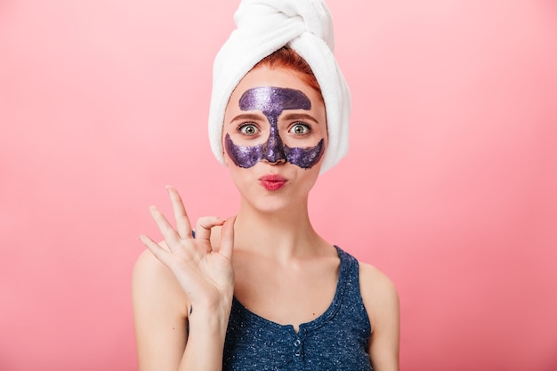Front view of woman with face mask showing okay sign. studio shot of amazed girl with towel on head gesturing on pink background. Free Photo