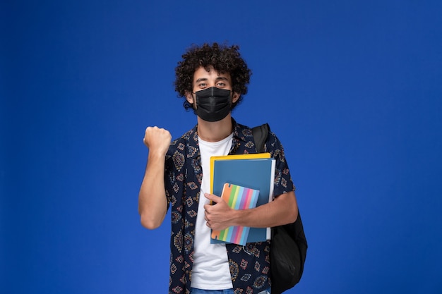 正面図水色の背景にコピーブックとファイルを保持しているバックパックと黒いマスクを身に着けている若い男子学生 無料の写真