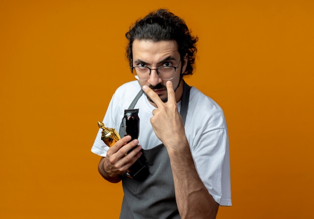 Frowning young caucasian male barber wearing glasses and wavy hair band in uniform holding winner cup and hair clippers doing i'm watching you gesture Free Photo