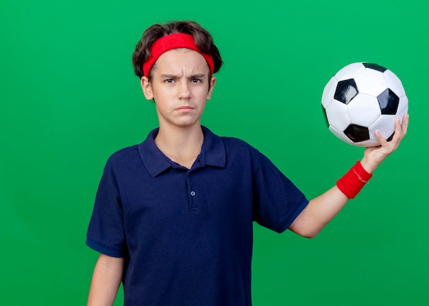 緑の壁に隔離された正面を見てサッカーボールを保持している歯科用ブレースとヘッドバンドとリストバンドを身に着けている若いハンサムなスポーティな少年をしかめっ面 無料の写真