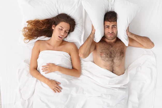 欲求不満の男は枕で耳を覆い 大声で妻がいびきをかくために眠ることができず 不眠症に苦しみ 寝室でポーズをとる いびきをかく女性にイライラする疲れた男 人 健康 睡眠障害 無料の写真