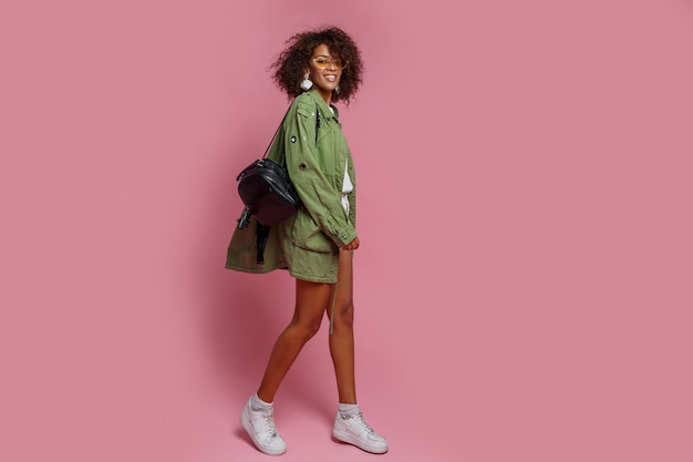 ピンクの背景のスタイリッシュな緑のジャケットに褐色肌の格好の良い女性の完全な長さの画像 ショッピングとファッションのコンセプトです 無料の写真