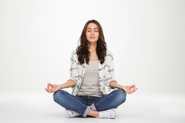 Full-length picture of concentrated woman in casual clothes meditating with closed eyes while sitting in lotus pose on the floor, isolated over white wall Free Photo